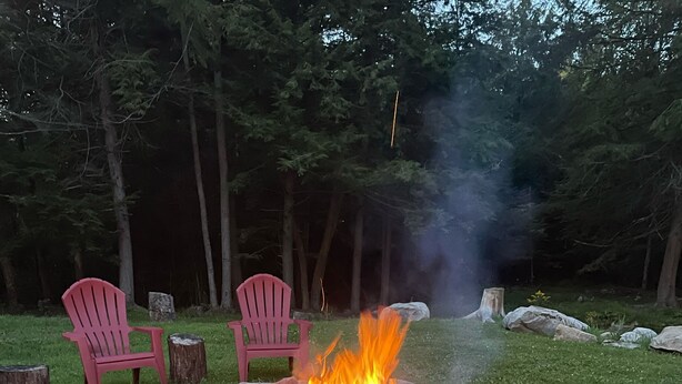 A fire pit with logs, surrounded by rocks and gravel, with two red chairs and a tree stump nearby. - Roscoe Cottage Pet Friendly  (Roscoe)