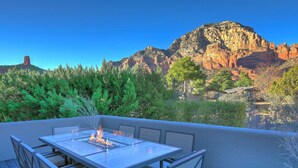 Un patio con mesa y sillas de vidrio, un elemento decorativo de fuego, y vistas a formaciones rocosas rojas y árboles.