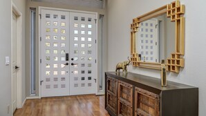 Un pasillo con piso de madera, una puerta blanca con paneles de vidrio y una consola de madera oscura con un espejo y objetos decorativos.