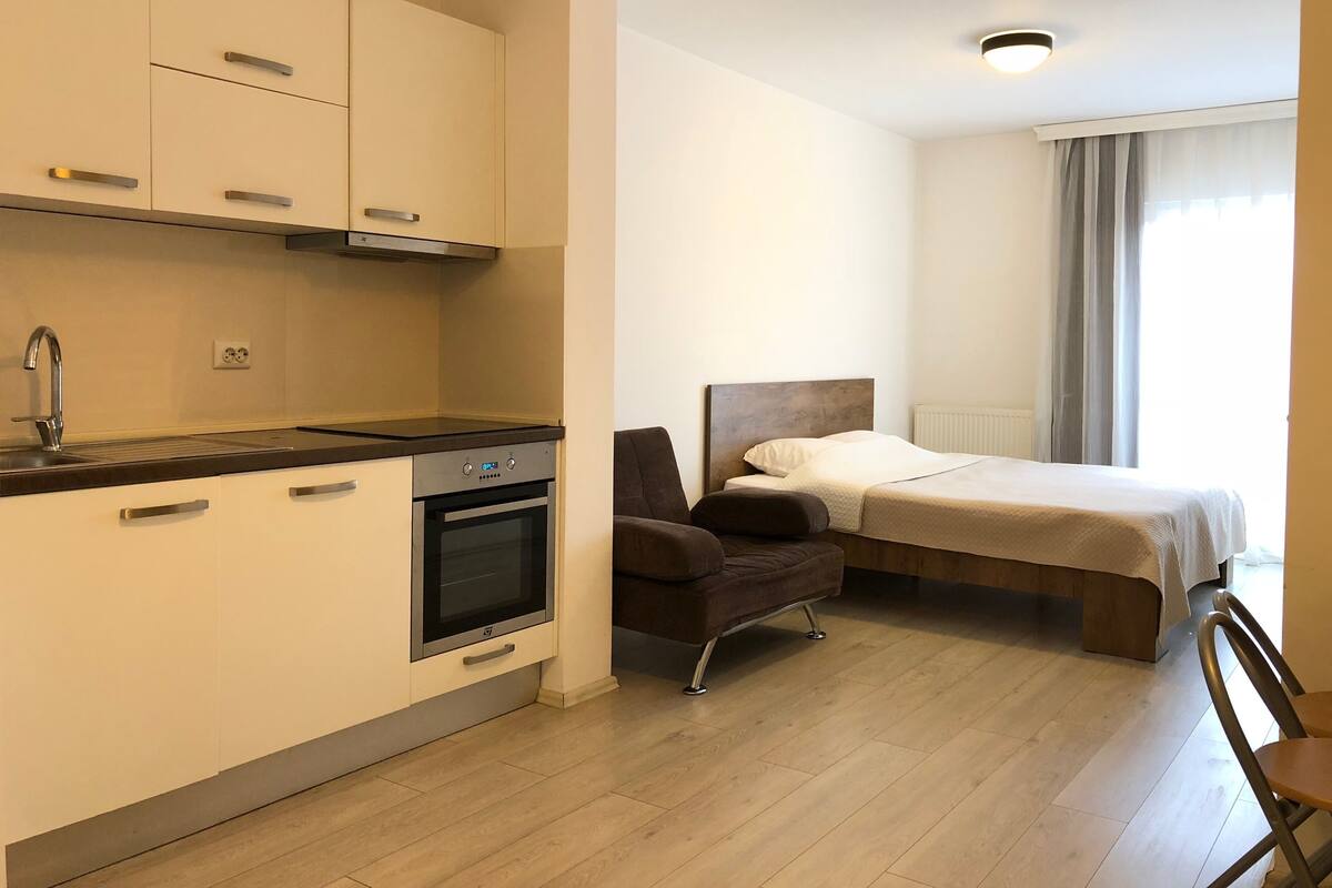 A compact hotel room with a kitchenette, a bed, a chair, and a view of the outdoors.