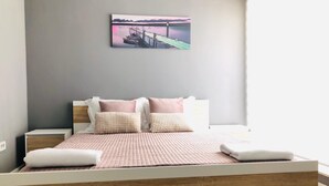 A modern bedroom with a wooden headboard, a large bed with pink bedding, and a view of a dock and water through a window. - Downtown Apartments (Pristina)