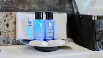 Uma bancada de banheiro com duas garrafas de HOLA BOLIGNA SHAMPOO & SHOWER GEL, um saboneteiro branco e uma bolsa preta.