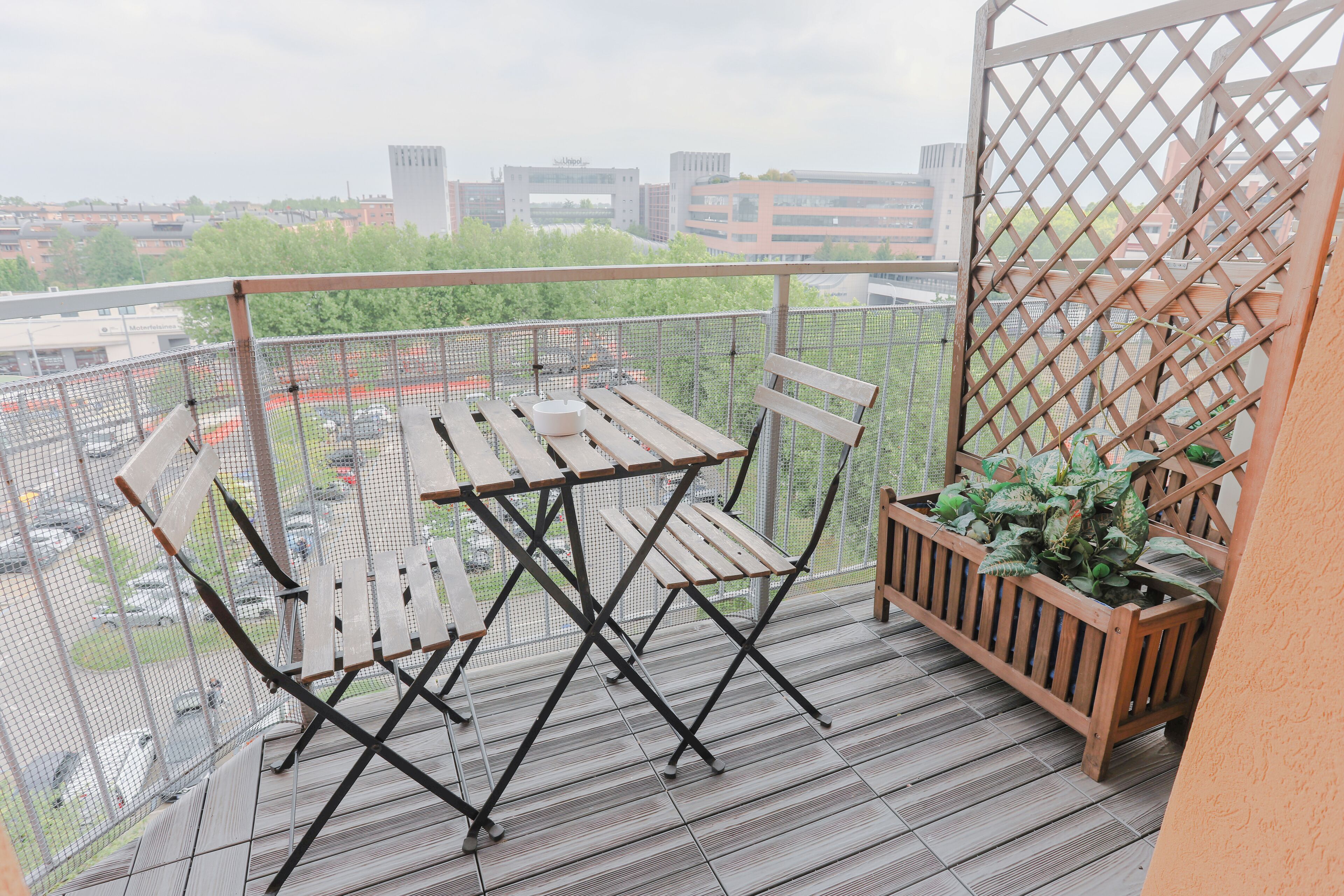 A balcony with a wooden table and chairs, a potted plant, and a view of the cityscape.