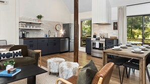 A modern kitchen with a dining area, featuring a wooden table set for four, a sofa with a fur throw, and a large window with a view of greenery.