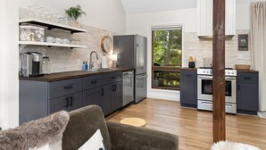 A modern kitchen with grey cabinets, stainless steel appliances, and a wooden countertop. There is a dining area with a grey sofa and a small round table. The kitchen has a large window with a view of greenery outside.