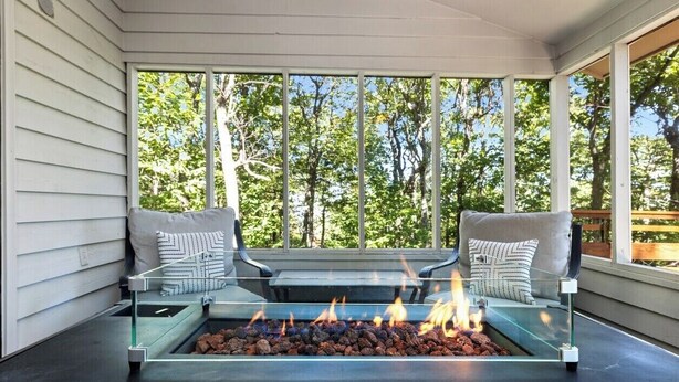 A modern outdoor seating area with a glass-top fire pit, two cushioned benches with pillows, and a view of trees through large windows.