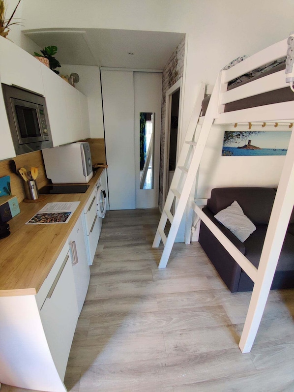 A compact kitchen with a microwave, toaster, and wooden counter. A small sofa with a cushion is positioned under a bunk bed with a painting above it.