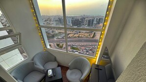 A high-rise room with a large window overlooking a cityscape, two grey armchairs, a small table with a remote control, and a television. - Heartland Hostel (Dubai)