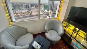 A room with a large window offering a city view, two grey armchairs, a small table with a remote control, and a TV on a stand. - Heartland Hostel (Dubai)