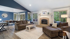Uma sala de estar com lareira, um espaço de jantar com mesa e cadeiras, e uma grande janela com vista para árvores.