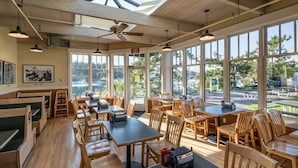 Restaurante com mesas e cadeiras de madeira, amplas janelas e vista para o exterior.