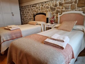 Ein Hotelzimmer mit zwei Betten, jedes mit einer beigen Decke und einem gefalteten Handtuch. Im Hintergrund der Betten eine Steinwand, ein weißer Kleiderschrank und ein Nachttisch mit Lampe und einer Dekoration.
