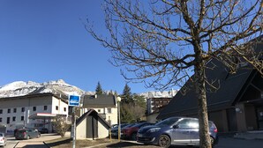 Un estacionamiento con coches, una señal de parada de autobús y edificios al fondo, con vista a la montaña.