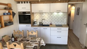Una cocina compacta con gabinetes blancos, horno empotrado y cafetera. Hay una mesa de comedor con mantel estampado y sillas de madera.