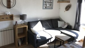 Una sala de estar acogedora con un sofá gris oscuro, una mesita de madera con una lámpara, un radiador y un cartel en la pared.