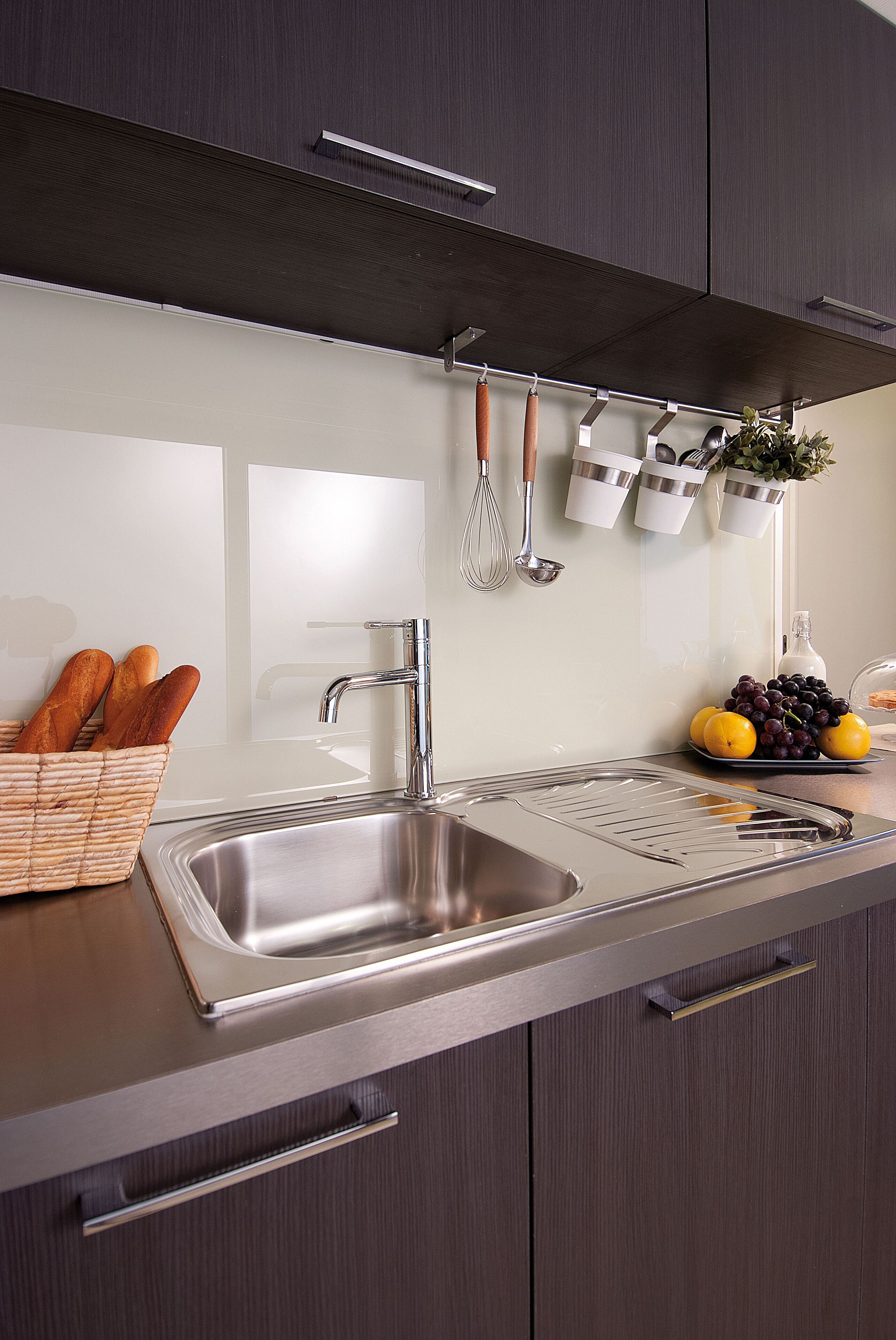 A modern kitchen with a stainless steel sink, a basket of baguettes, and a fruit bowl with lemons and grapes.