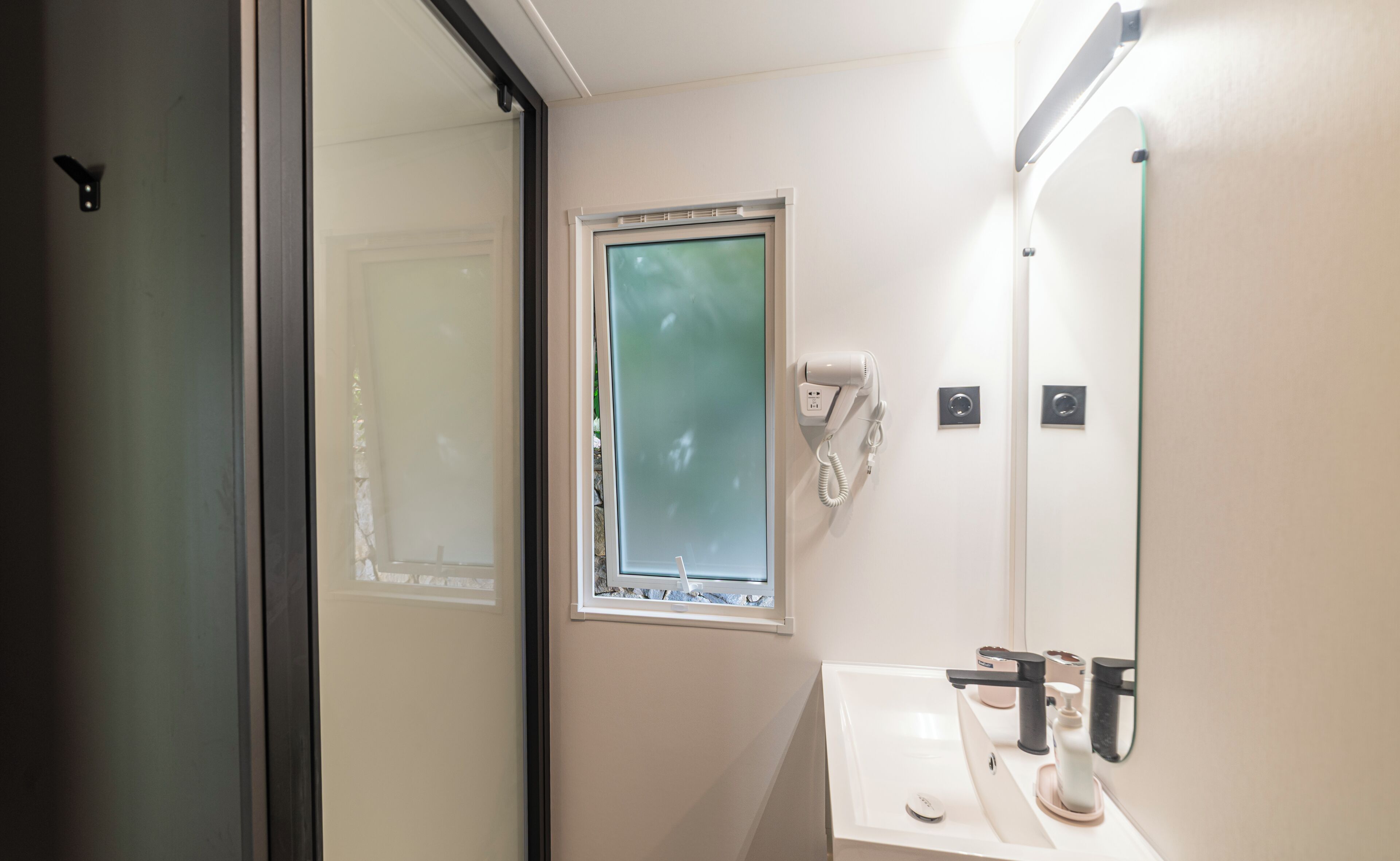 Ein Badezimmer mit Glastür, Fenster, Fön und Waschbecken mit Spiegel.