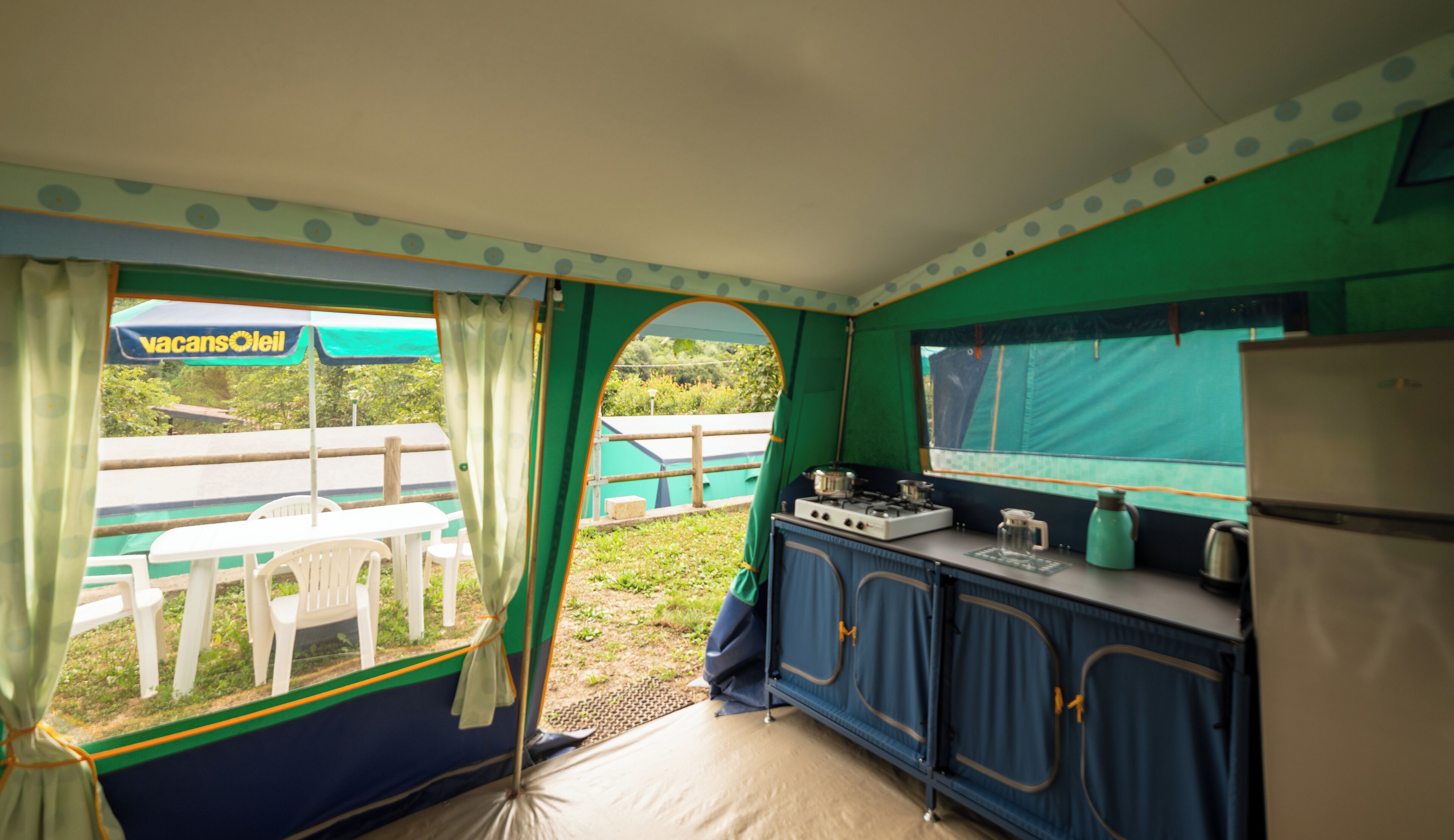 Cocina compacta con refrigerador, estufa y lavamanos. Se ve un área de comedor con una mesa y sillas a través del ventanal.