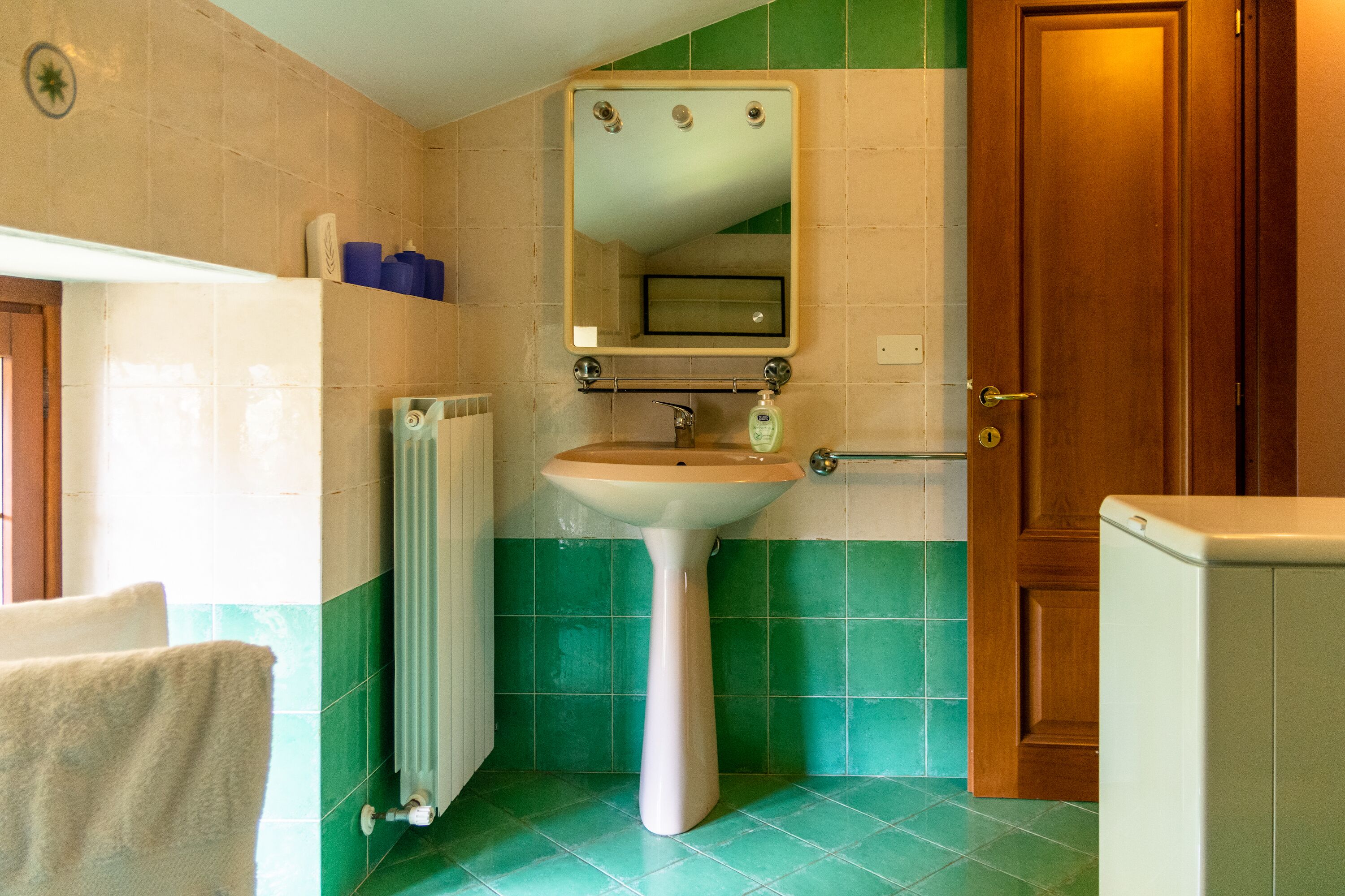 Un baño con un lavamanos blanco de pedestal, un espejo encima, una toalla colgada en la pared y una puerta de madera.