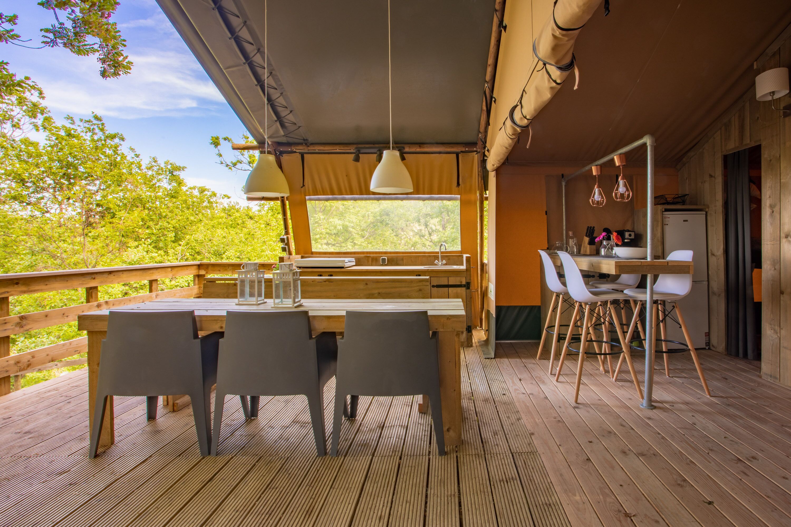 Een houten terras met een tafel en stoelen, een keuken met een koelkast, en een groot raam met uitzicht op groen.