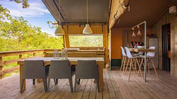 Een houten terras met een tafel en stoelen, een keuken met een koelkast, en een groot raam met uitzicht op groen.