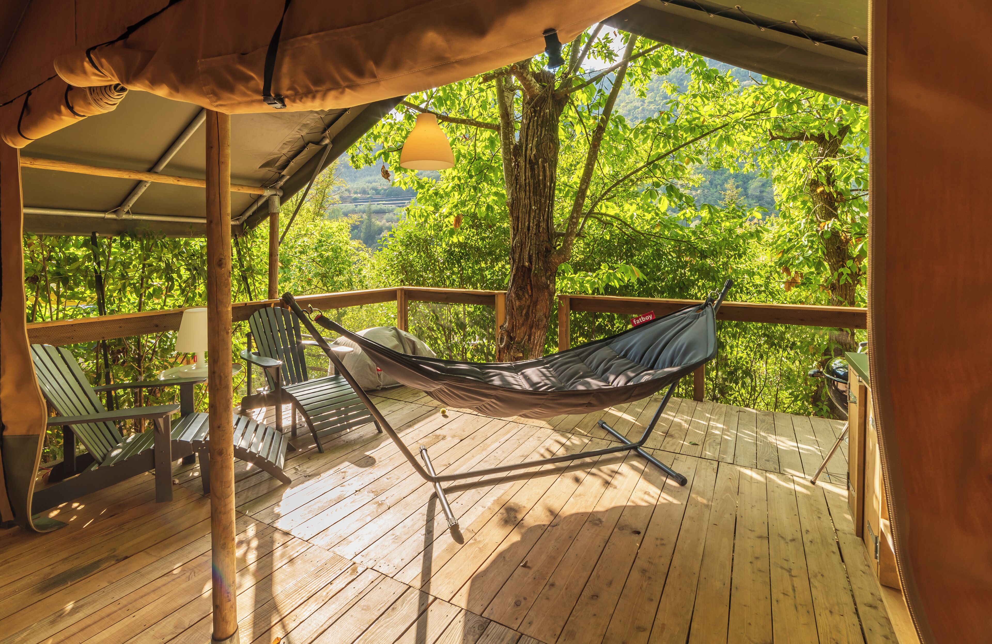 Un hamac sur une terrasse en bois, agrémentée de chaises, le tout entouré d’une végétation luxuriante.