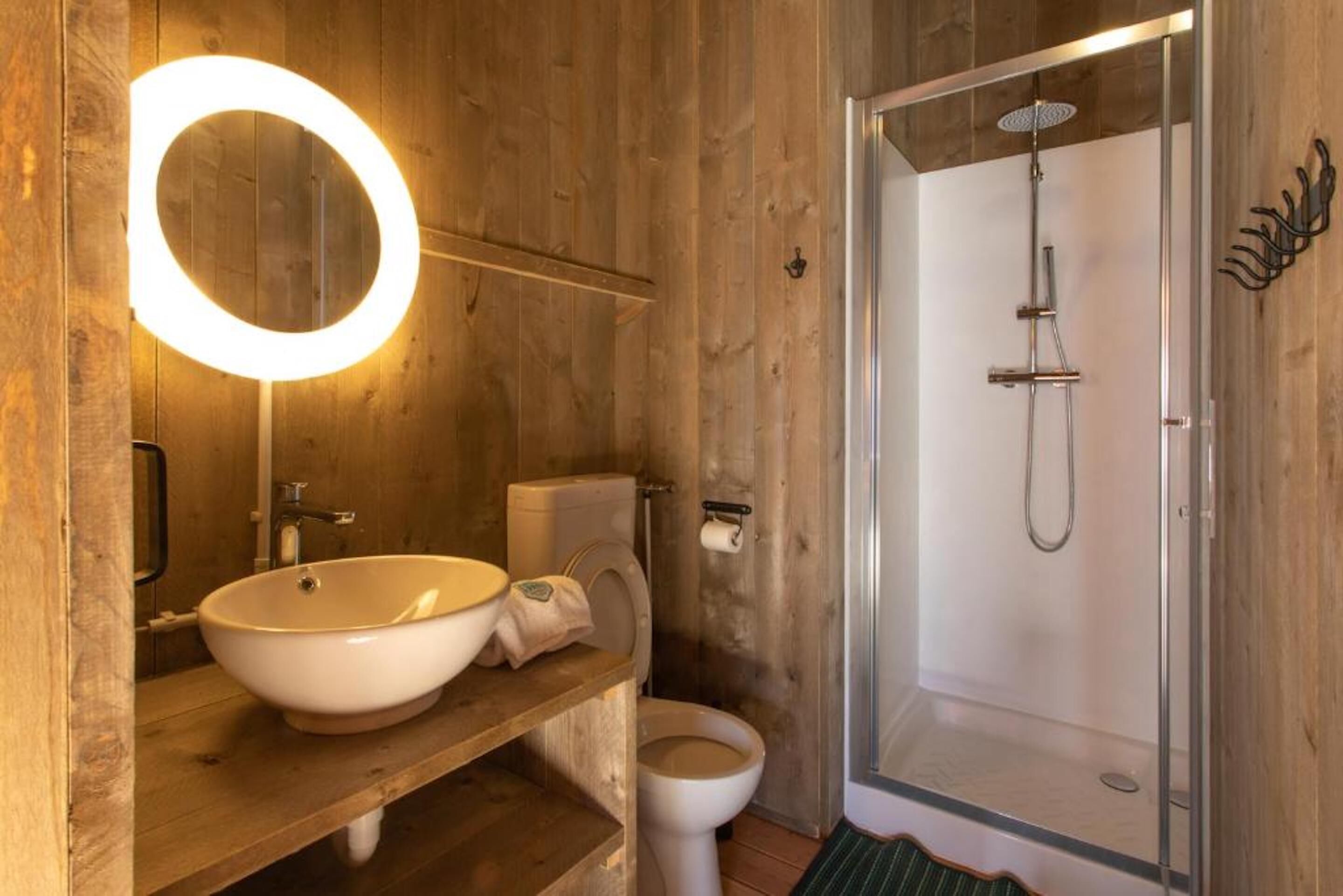 Ein Badezimmer aus Holz mit einem runden Spiegel, einem Waschbecken, einer Toilette und einer Duschkabine.