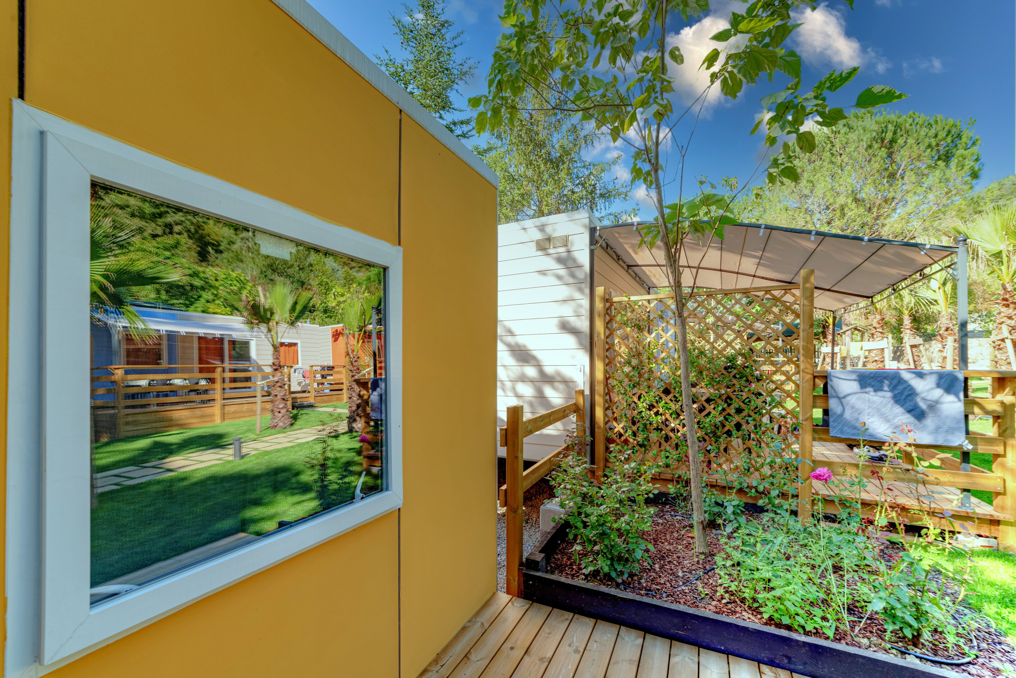 Ein gelbes Gebäude mit einem Fenster, einer Holzterrasse und einem Gartenbereich mit Pflanzen und einem Holzzaun.