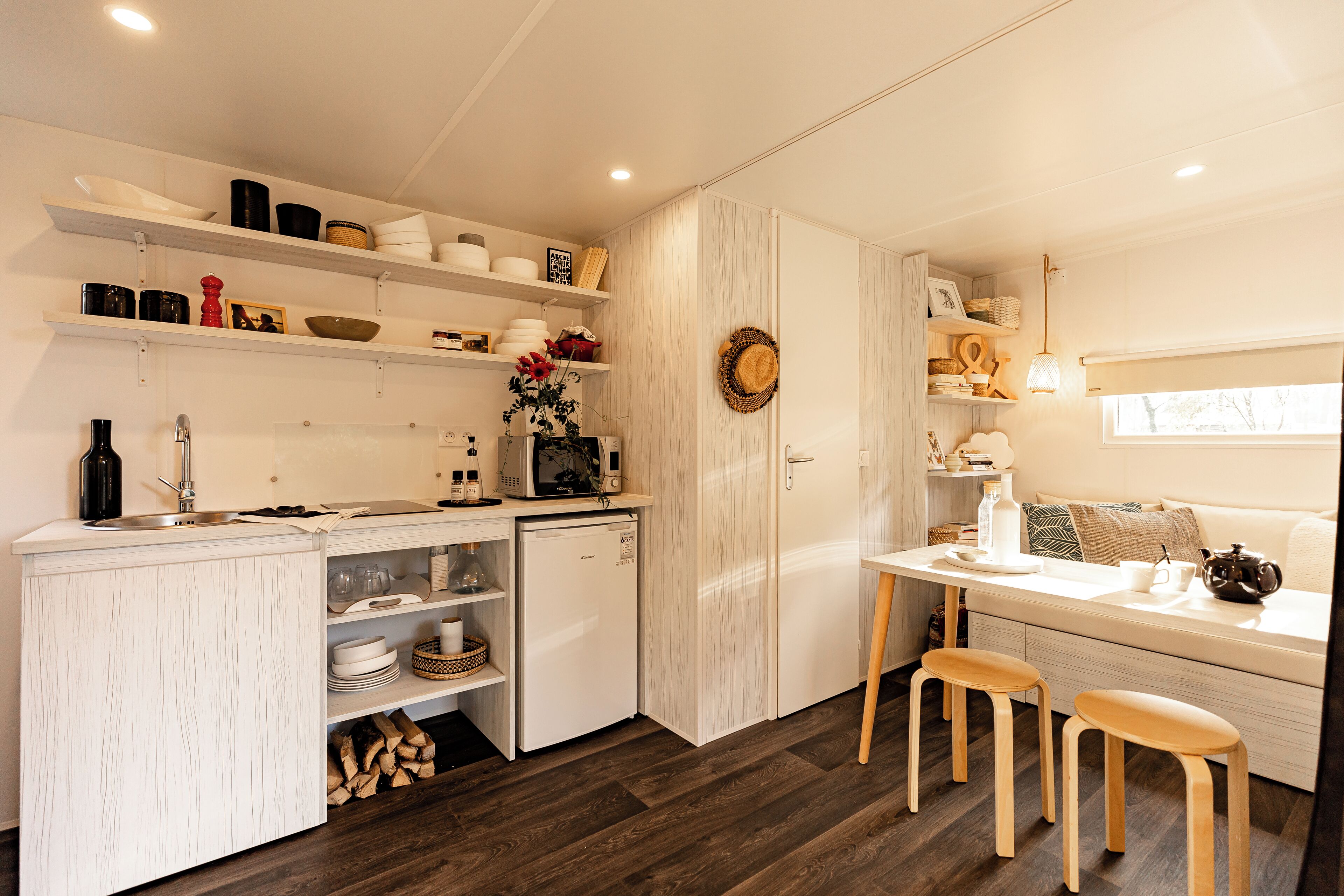 A compact kitchen with white cabinetry, a built-in microwave, and a small refrigerator. There are wooden stools at a small dining table, and a window with a view of the outdoors.