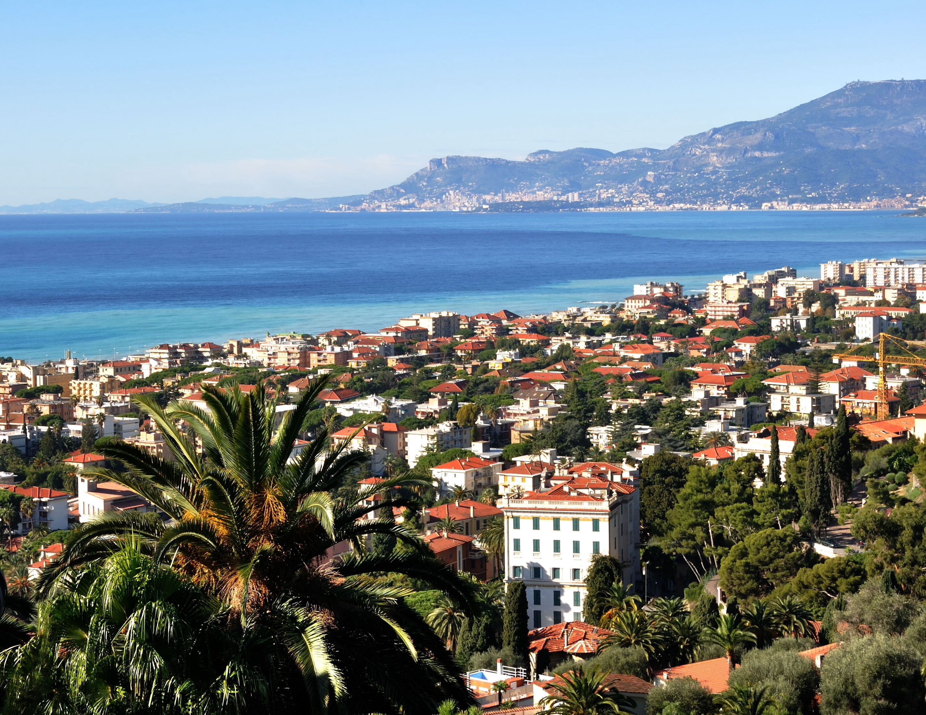 A coastal cityscape with dense urban buildings, a prominent white building with a red roof, and a clear view of the sea and distant mountains.