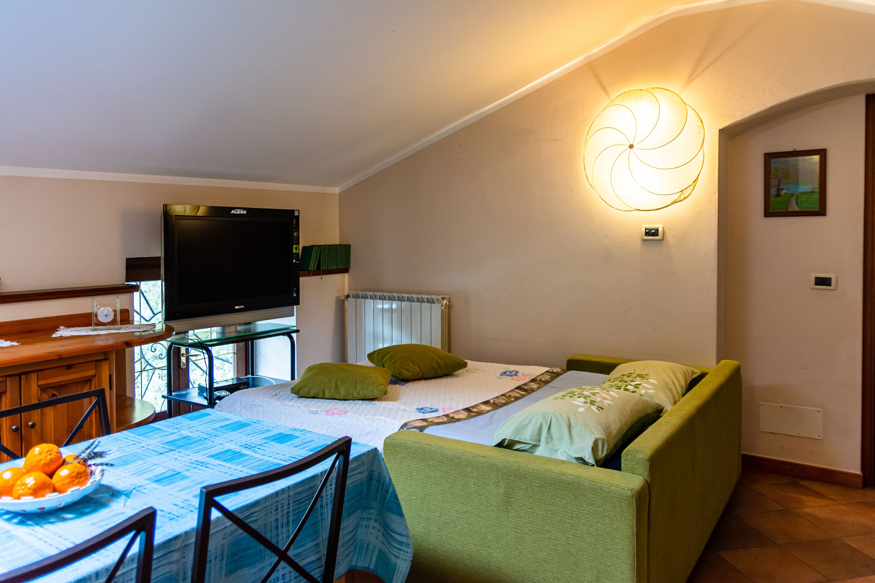 A cozy room with a bed, a television, a dining table with chairs, and a bowl of fruit.