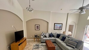Uma sala de estar com sofá seccional, televisão sobre suporte de madeira, mesa de centro de vidro e tapete estampado.