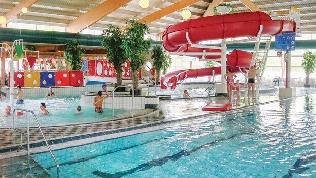 Sisätiloissa sijaitseva uima-allas, jossa on useita uimaratja, vesiliukumäki ja värikkäillä rakenteilla varustettu leikkialue.