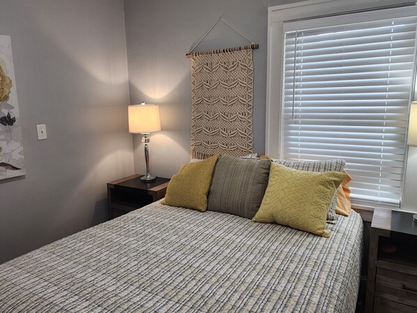 Un dormitorio con cama, lámpara de noche, cortina estampada y ventana con persianas.