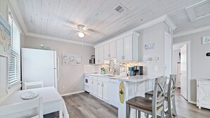 A kitchen with white cabinetry, a central island, and a dining area with white chairs. - Super Nice Quiet Beach Cottage, Pvt Parking, WiFi, Cozy, Spotless, Comfortable.  (Gulf Shores)