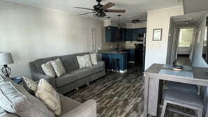 A modern living room with a grey sofa, wooden flooring, and a kitchen area with blue cabinets. - 1st Floor Bayfront 1 bedroom Updated Condo with pool overlooking the bay!! (Ocean City)