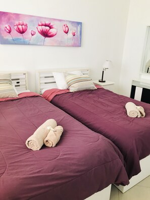 A hotel room with two beds, each with a purple bedspread and rolled towels. There is a painting of pink flowers on the wall above the beds.
