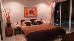 Une chambre avec un grand lit, une table de chevet avec une lampe, un vase contenant de grandes plantes et un tableau représentant des fleurs accroché au mur.