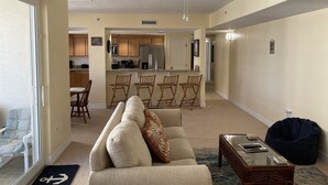 Un salón con un sofá, una mesa de centro y un sillón de granos de espuma. Se ve una cocina al fondo con un refrigerador, estufa y sillas.