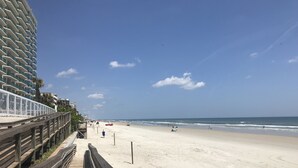 Una playa con un paseo marítimo de madera, cielo despejado y vista distante del océano.