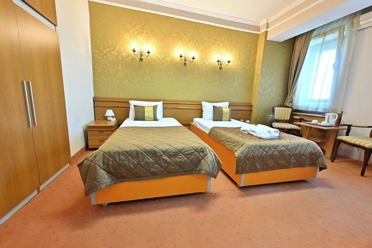 A hotel room with two beds, a wooden wardrobe, a nightstand, a lamp, and a window with curtains.