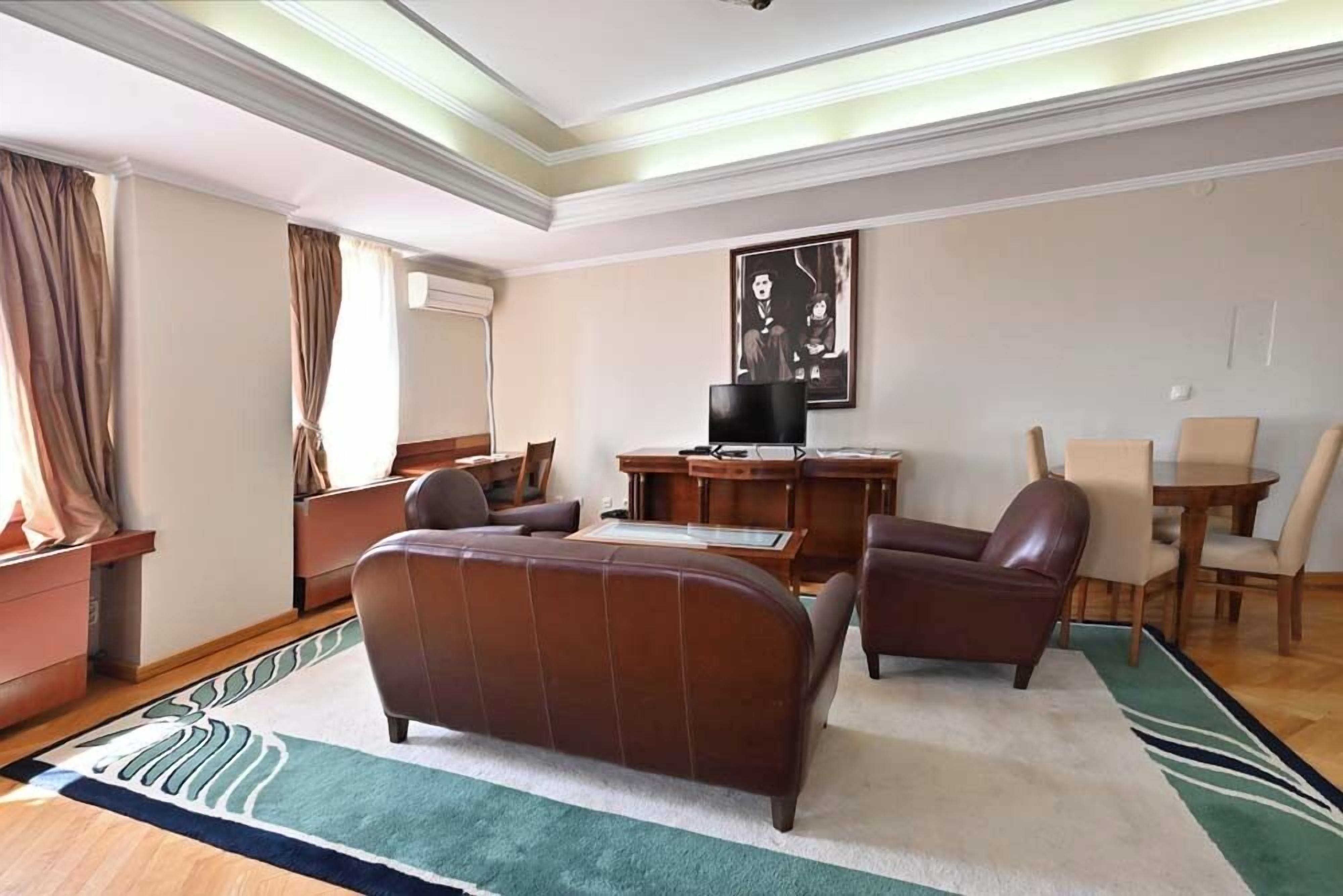 A hotel room with a large brown leather sofa, a wooden desk, a television, and a dining area with chairs.