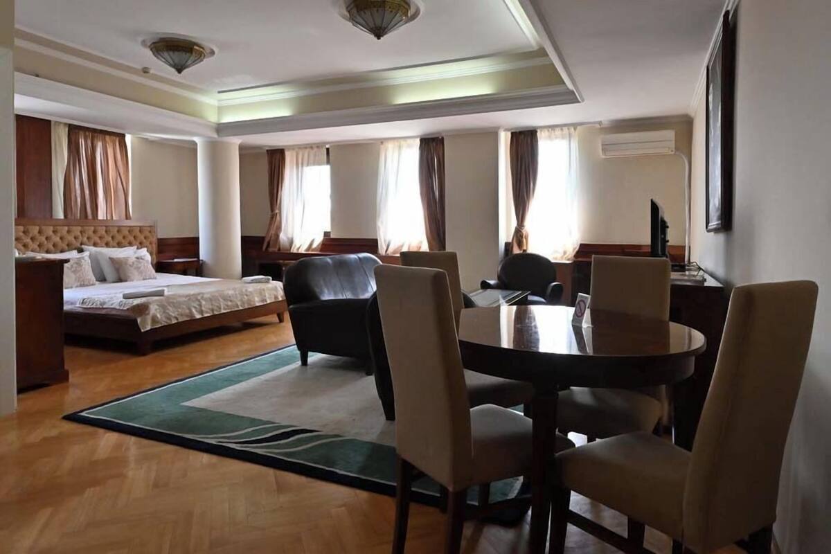 A hotel room with a bed, a desk, a dining table with chairs, and a large window with curtains.