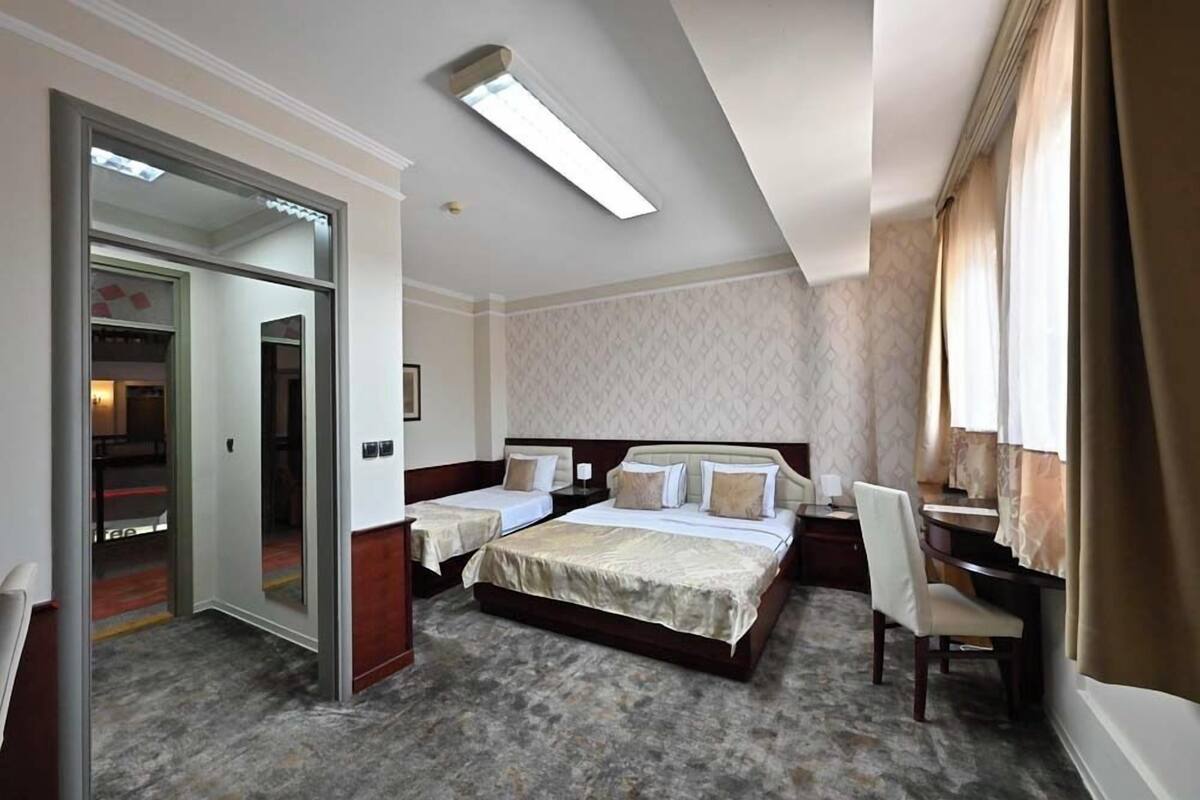 A hotel room with a large bed, a desk, and a chair. There is a wardrobe and a mirror.
