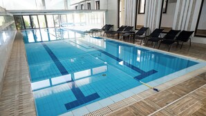 A large indoor swimming pool with clear blue tiles, wooden flooring, and several black lounge chairs along the poolside. - Hotel Sole Mio (Novi Sad)