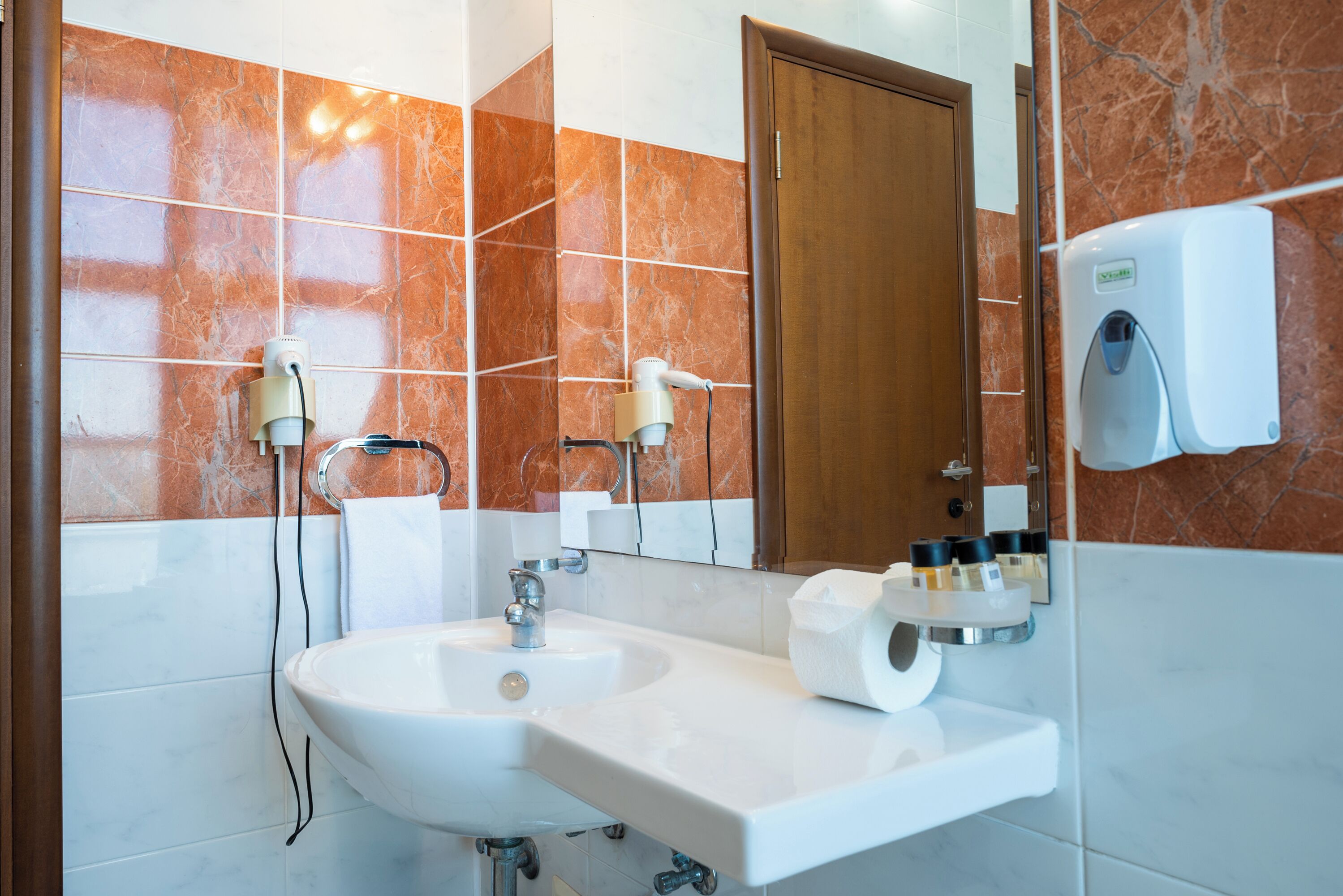 A modern bathroom with a white sink, two hair dryers, a towel dispenser, and a mirror.