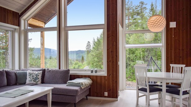 A cozy room with a sofa, a small table, and a dining area with chairs. Large windows offer a view of the forest and mountains. - Cozy home in Skollenborg with kitchen (Skollenborg)