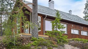 A wooden cabin with large windows, a chimney, and a deck area surrounded by trees and shrubs. - Cozy home in Skollenborg with kitchen (Skollenborg)