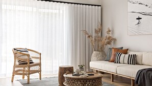 Un salon comprenant un fauteuil en osier, une table basse ronde en bois, un canapé avec des coussins et une grande fenêtre ornée de rideaux légers.