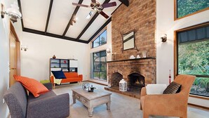 A living room with a fireplace, large windows, and a ceiling fan. - Serendipity Gardens! (Joyner)